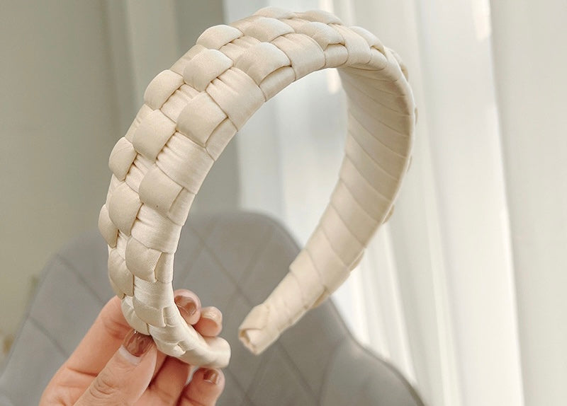 A woman holding the Beau headband, buttery ivory colour against a soft neutral background. Cross-hatched satin silk design, wrapped around the headband. Casual or formal addition to any event outfit, day or night.