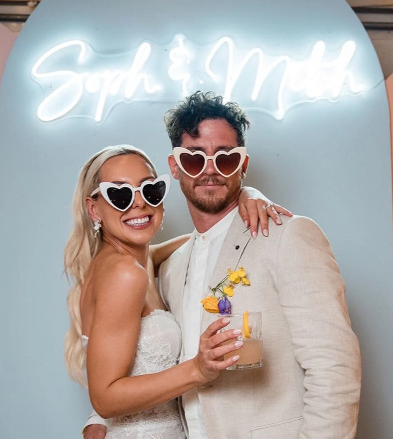 A Bride and Groom posing for a picture with our cream Heart shaped sunglasses on. With a backdrop of light pastel blue and neon name lettering behind them. Bride is wearing the white colour way and the Groom is wearing the Cream with hue lenses colour way. Colour comparisons and how they are perfect for weddings