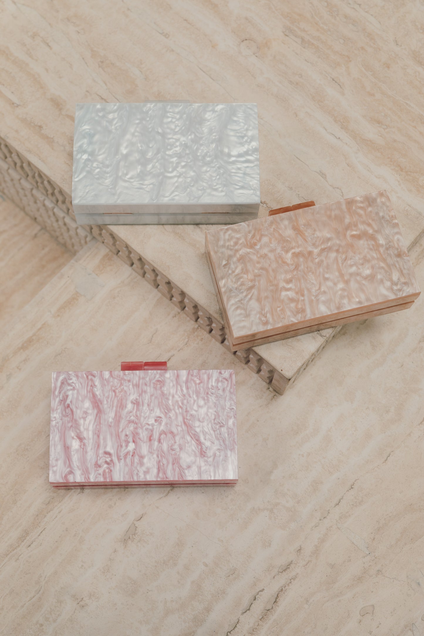 A shot of our 3 colour variations of our candy acrylic bag, shown with both straps removed and the soft marbled metallic sheen of each colour way. White at the top, apricot in the middle and pink at the bottom, against a neutral beige background. The perfect hand held clutch bag for and evening party or date night