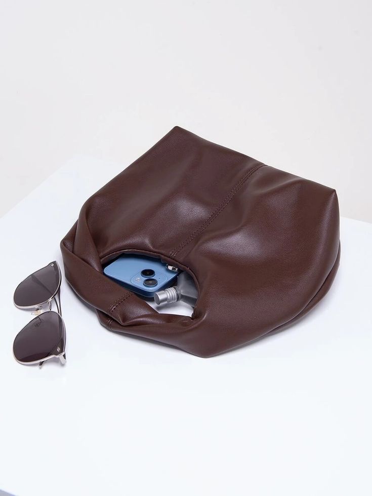 Our rich chocolate PU leather bag, laying flat on a white surface, showcasing the contents of what the bag can hold for size comparison. Inside the bag there is a smart phone, perfume, sunglasses and other essentials. Highlighting it's compact size and functionality. Perfect for day to night events or after work drinks, a smart and luxurious bag for all occasions