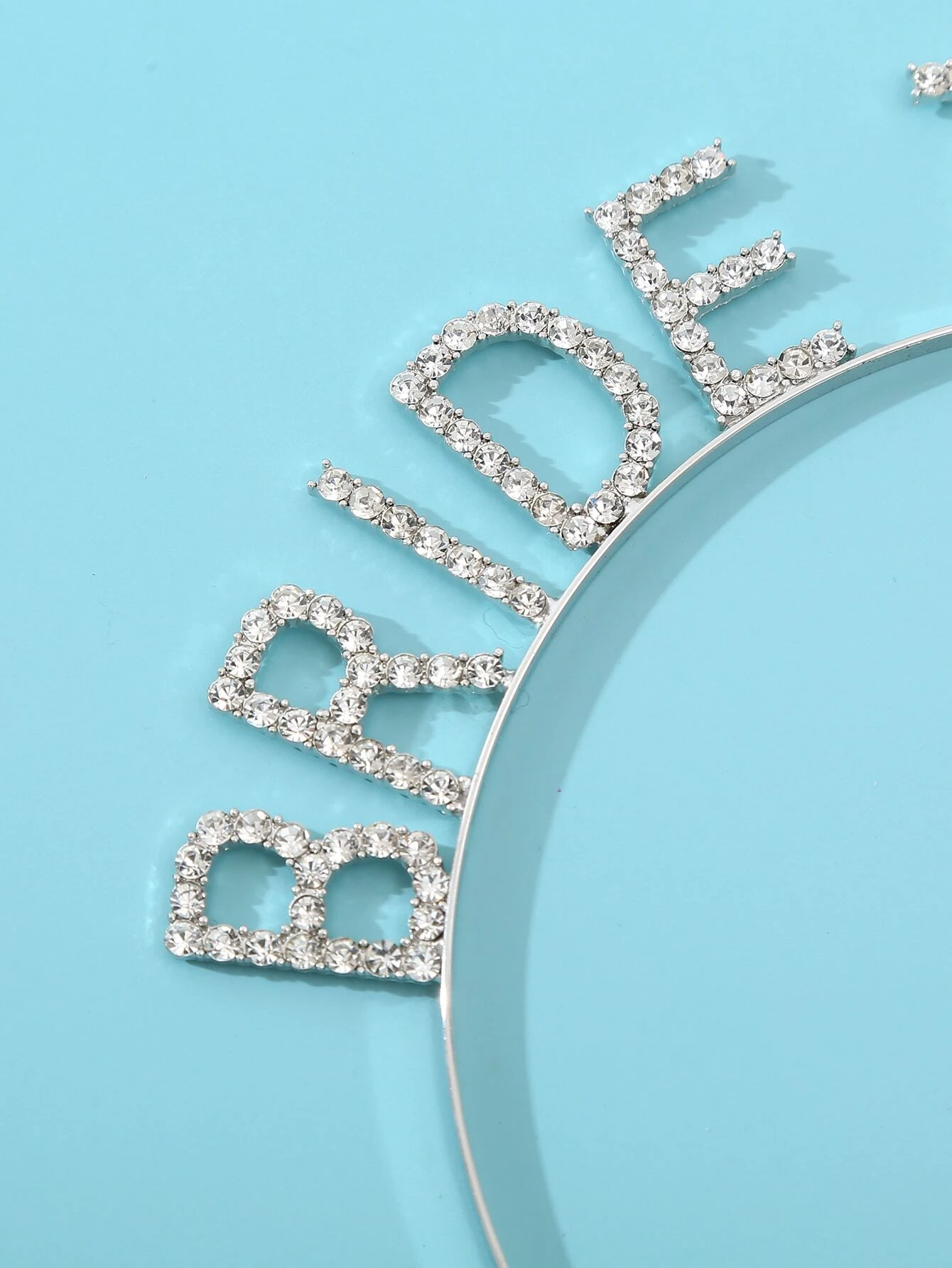 Close-up of the 'Bride' lettering in our 'Bride-to-be' Diamante Headband on a baby blue surface. Showing the sparkle from the diamante rhinestones lettering. Adding glamour to every brides pre wedding celebrations.