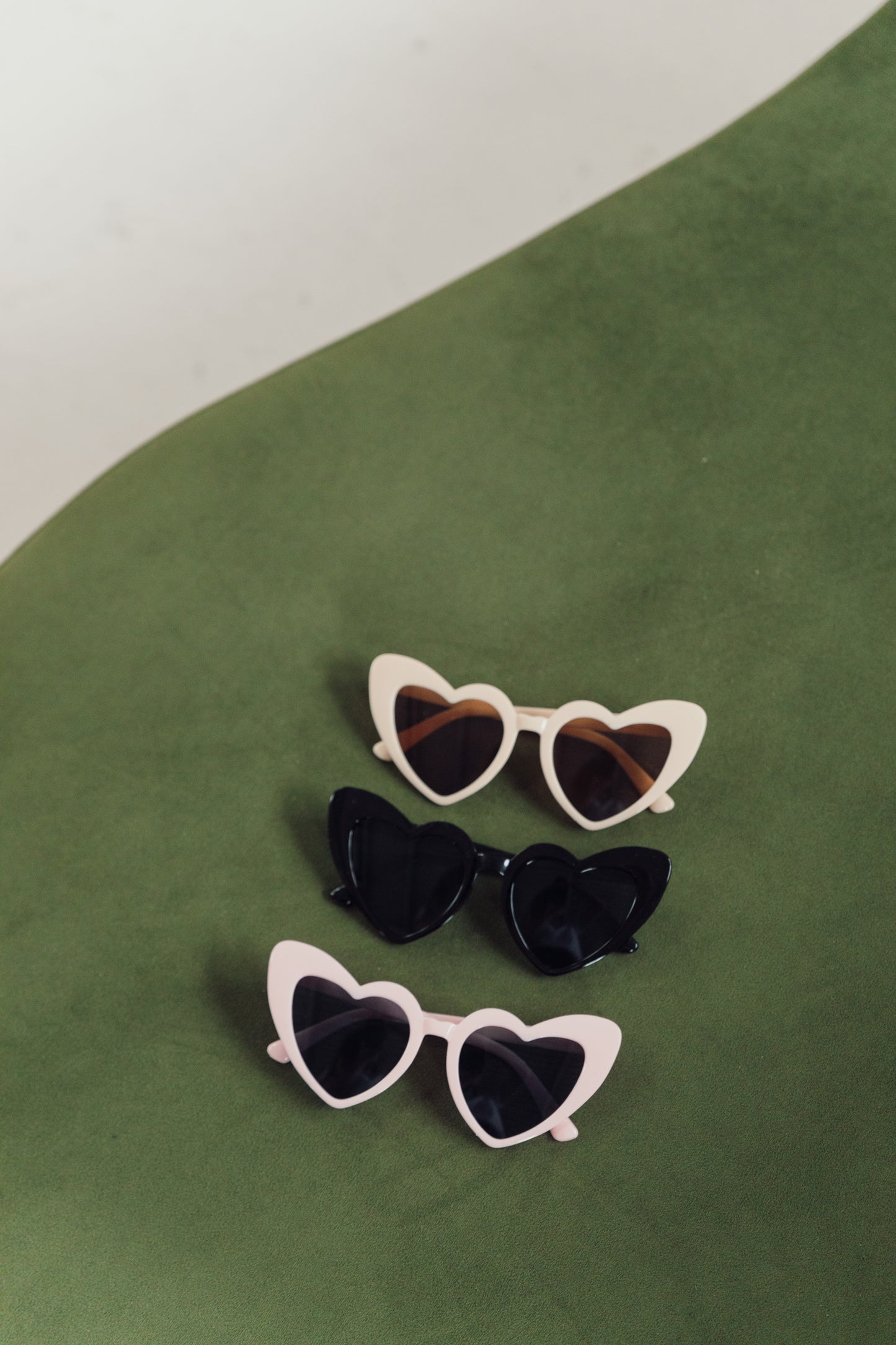 Olive green background with three heart shaped sunglasses laying flat. Cream at the top with a lense hue, black in the middle with solid blackout lense and pink and black at the bottom. Highlighting their fun shape and colour comparisons. Perfect for hen parties or theme nights