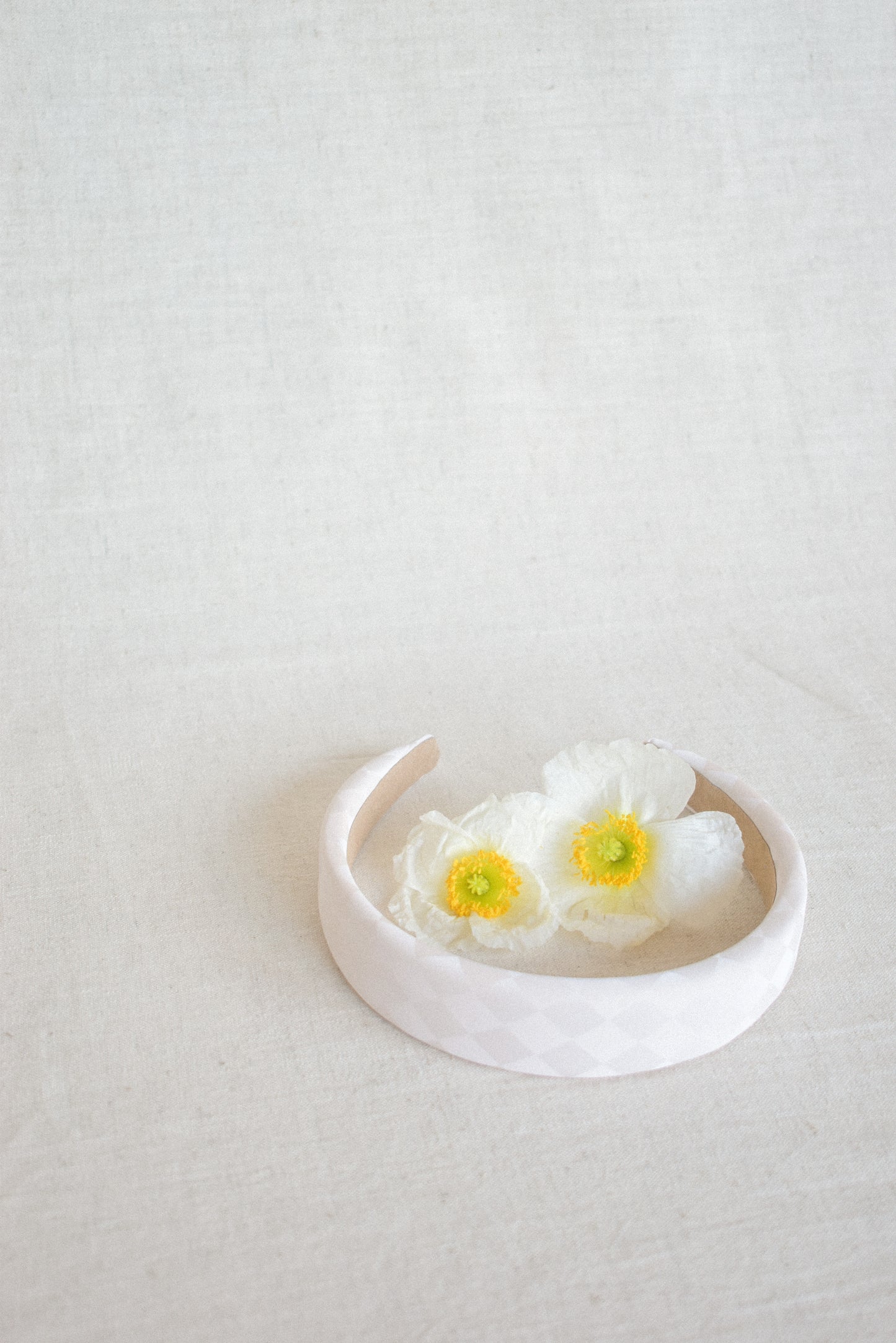 Our cream coloured Dahlia Headband laying flat on a white surface with white and yellow flowers placed in the middle of the band. Colours of the flower compliment the cream checked satin band, offering inspiration for outfit pairings. Spring races or bridal parties this would be perfect for