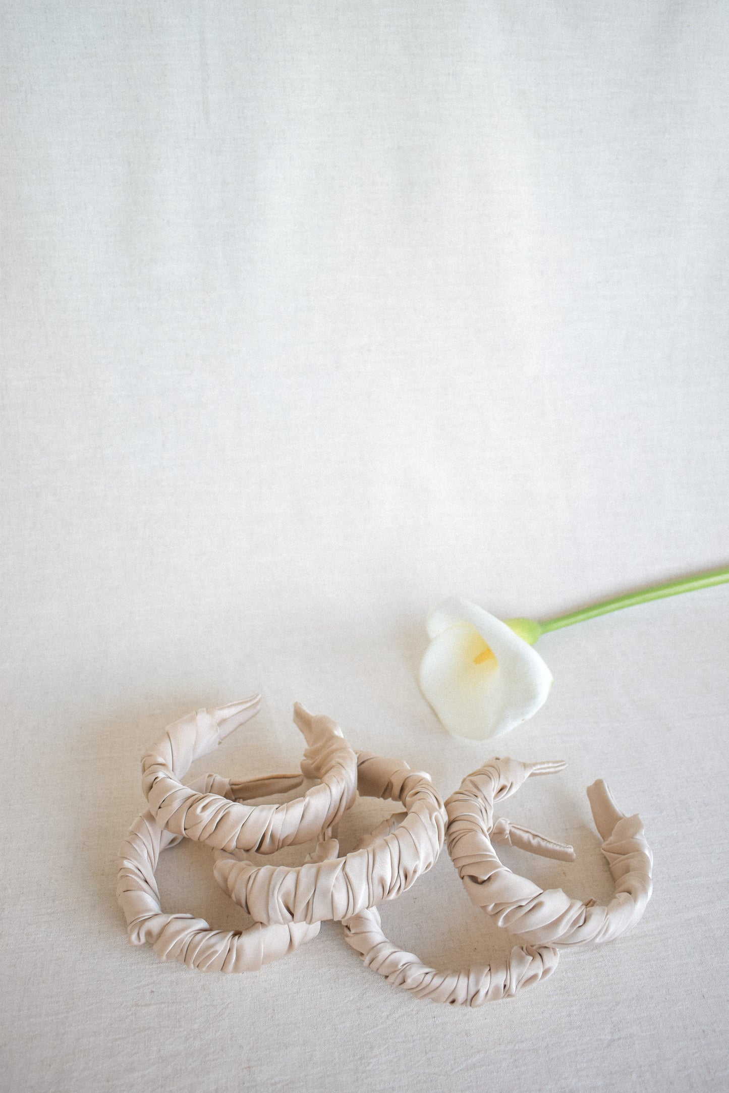 A scattered pile of our Cloud latte coloured headbands against a cream background and a peace lily flower. Ruched and wrapped satin/silk around the entire padded band. Adding elegance and class to any bridal party