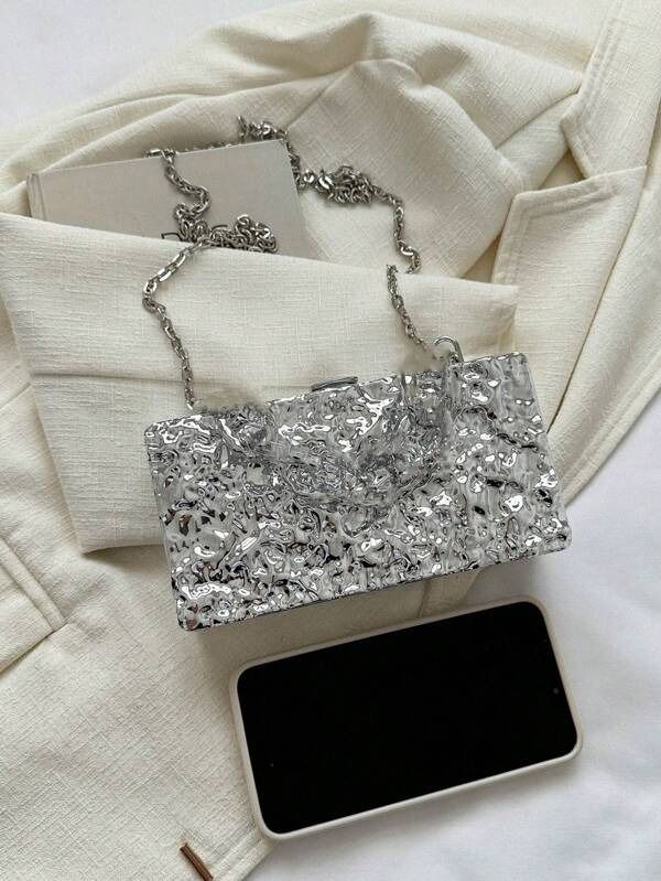 Our Lexi chrome mirrored bag laying flat on top of a textured neutral blazer, with the shoulder chain attachment. For size comparison, a smart phone is shown next to the bag, highlighting the compact size and practicality of the clutch bag for your next night out.