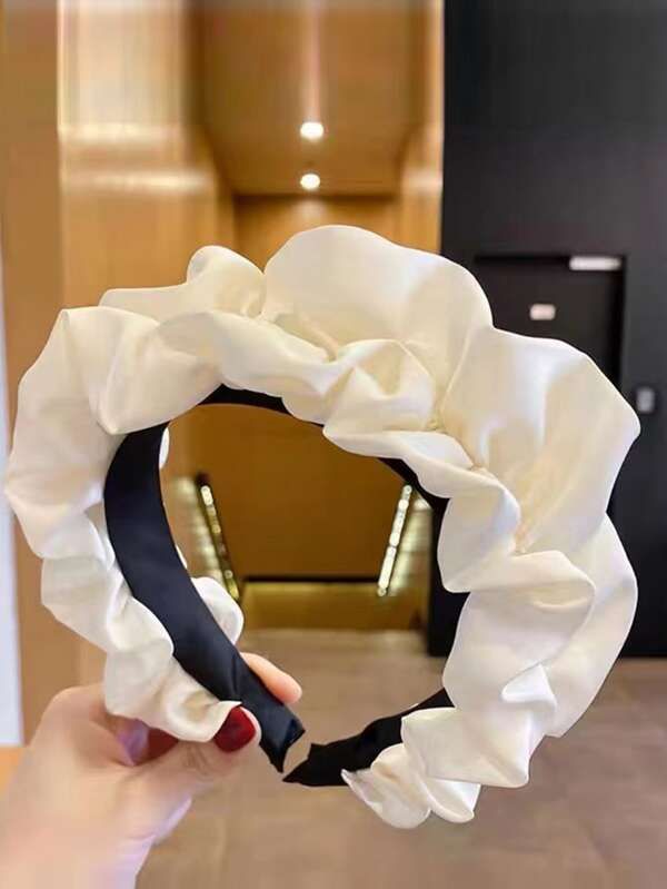 Our Lotus headband being held at an angle by a woman, showing a close up of the soft ruched design and cream silk/satin material across the whole band. Highlighting the band underneath the ruffles is a black satin, for comfort and blending. Perfect for brides/bridesmaids