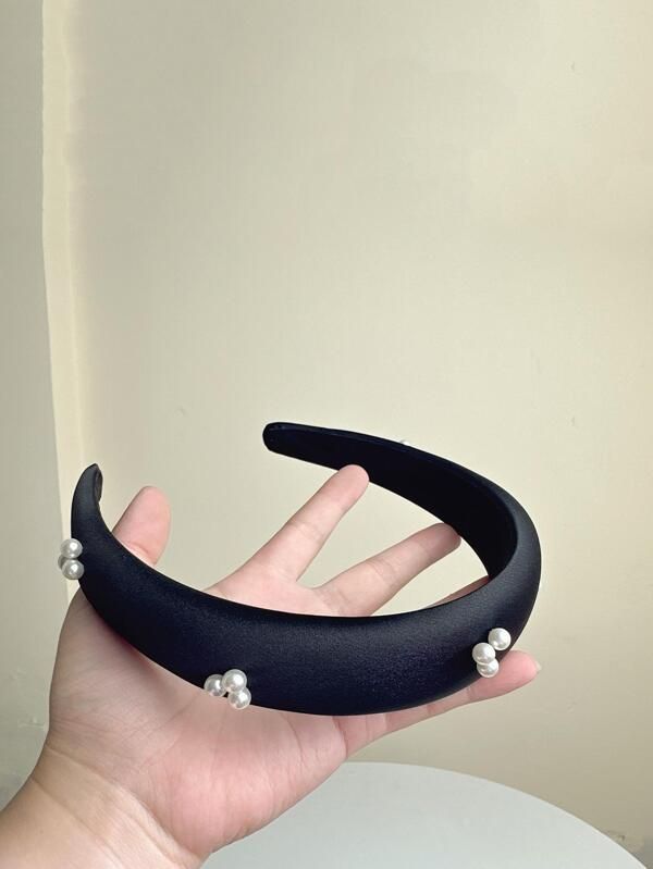 Our Noir headband being held flat in a hand, highlighting a size comparison and sheen of the black satin fabric with small pearls clustered and spaced out along the band. Perfect for formal events or daytime glamour