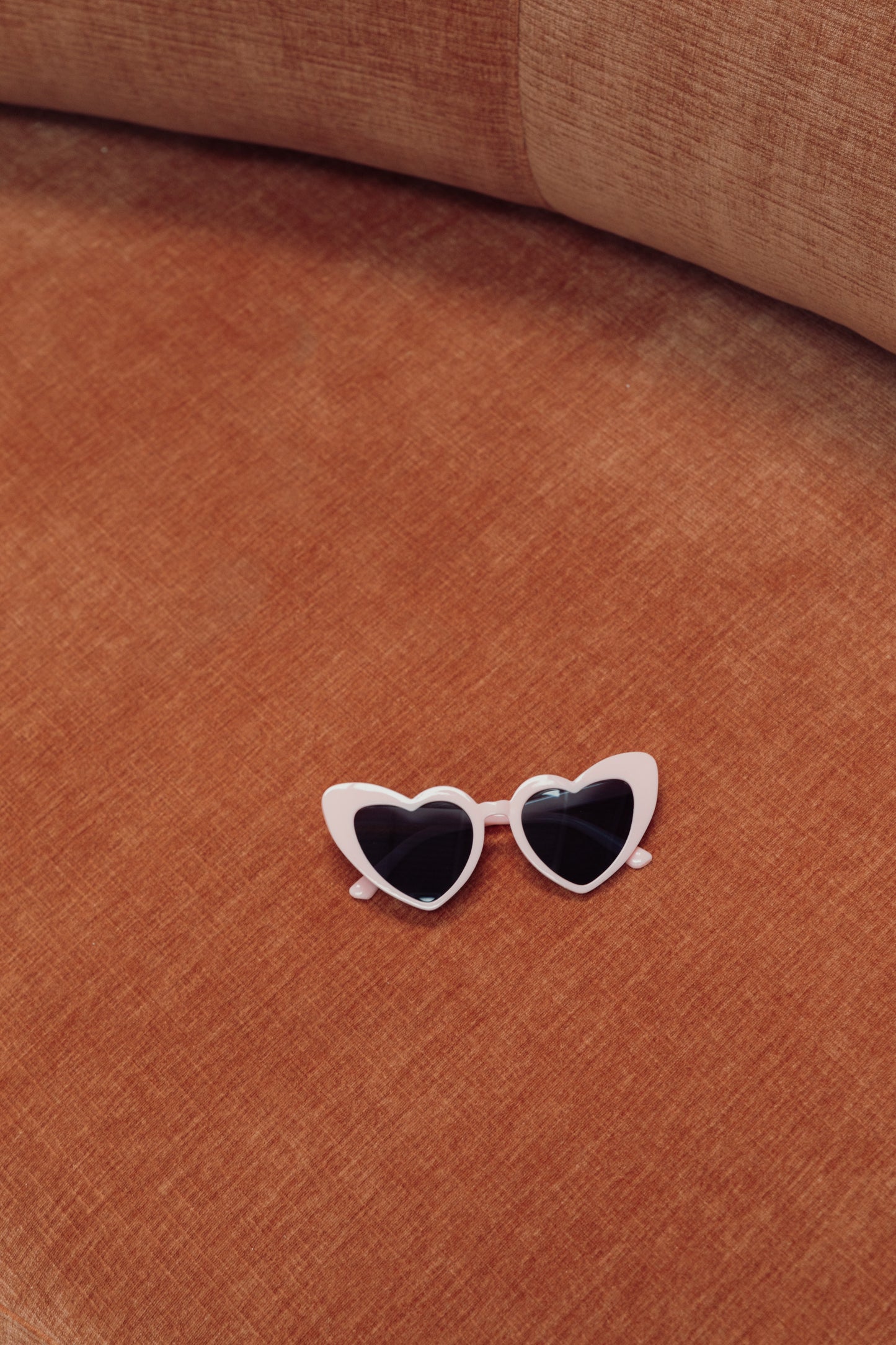 Our Pink on Black Heart Sunglasses, against a terracotta couch. These come in various colours, highlighting the pale pink colour with black lenses heart-shaped glasses. Perfect for bride or bridesmaids at your next hen party