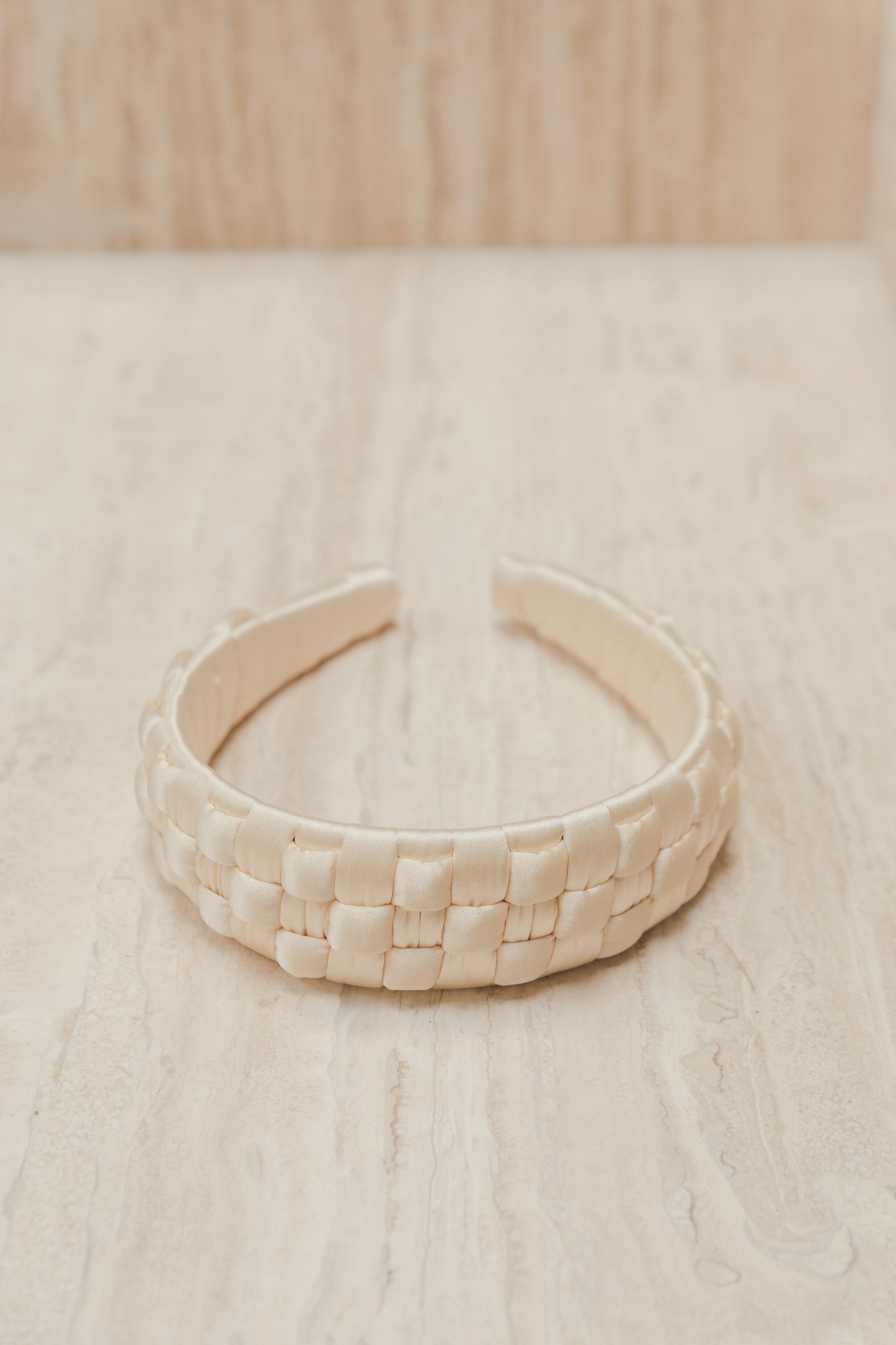 Our Beau headband shown laying flat on a marble surface. Buttery ivory in colour showing a different angle of the cross-hatched satin silk design, wrapped around the headband. Casual or formal addition to any event outfit, day or night.