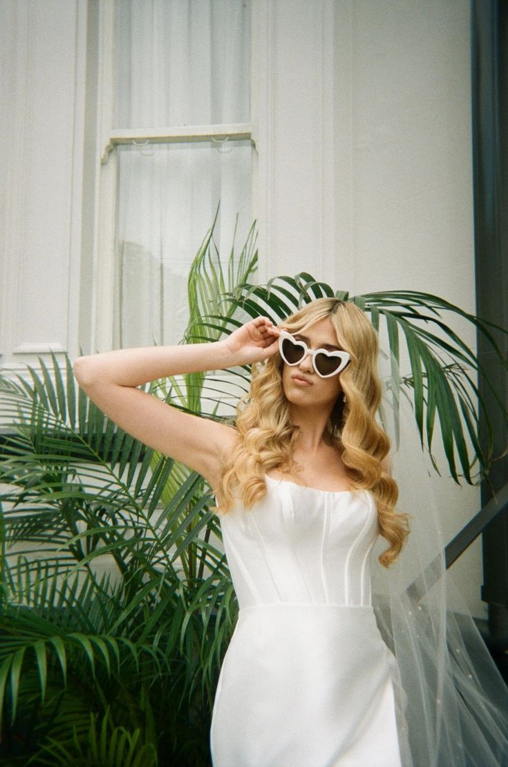 A bride wearing the cream heart sunglasses, holding one side in a fun pose with a palm in the background. Inspiration on how to style, perfect for bride to be or wedding day