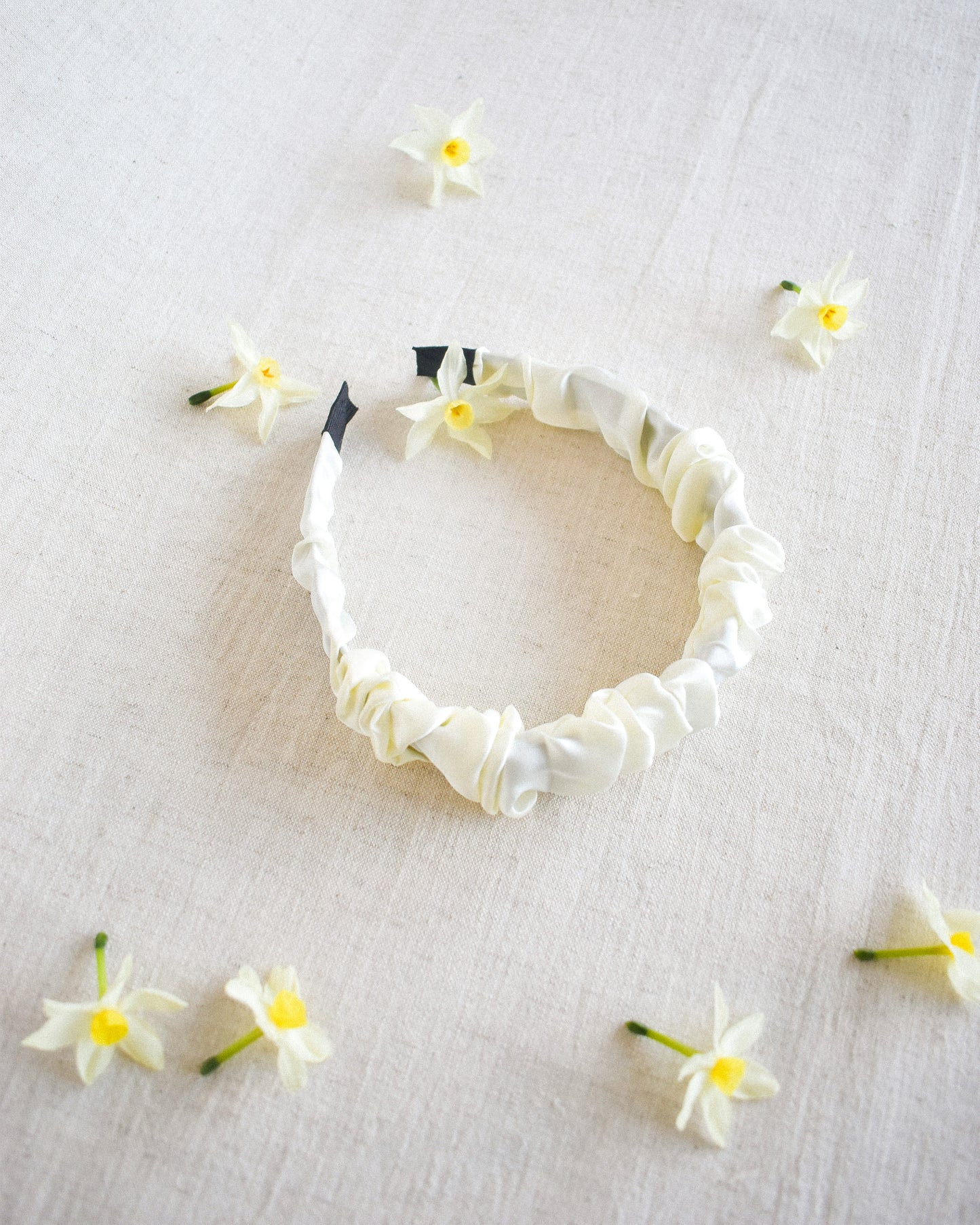 Our Calla headband shown laying down on a neutral linen surface with small white and yellow flowers scattered. Soft cream in colour, satin/silk material that is ruffled across the whole band. Perfect for bridal parties, spring season or dressing up a casual outfit