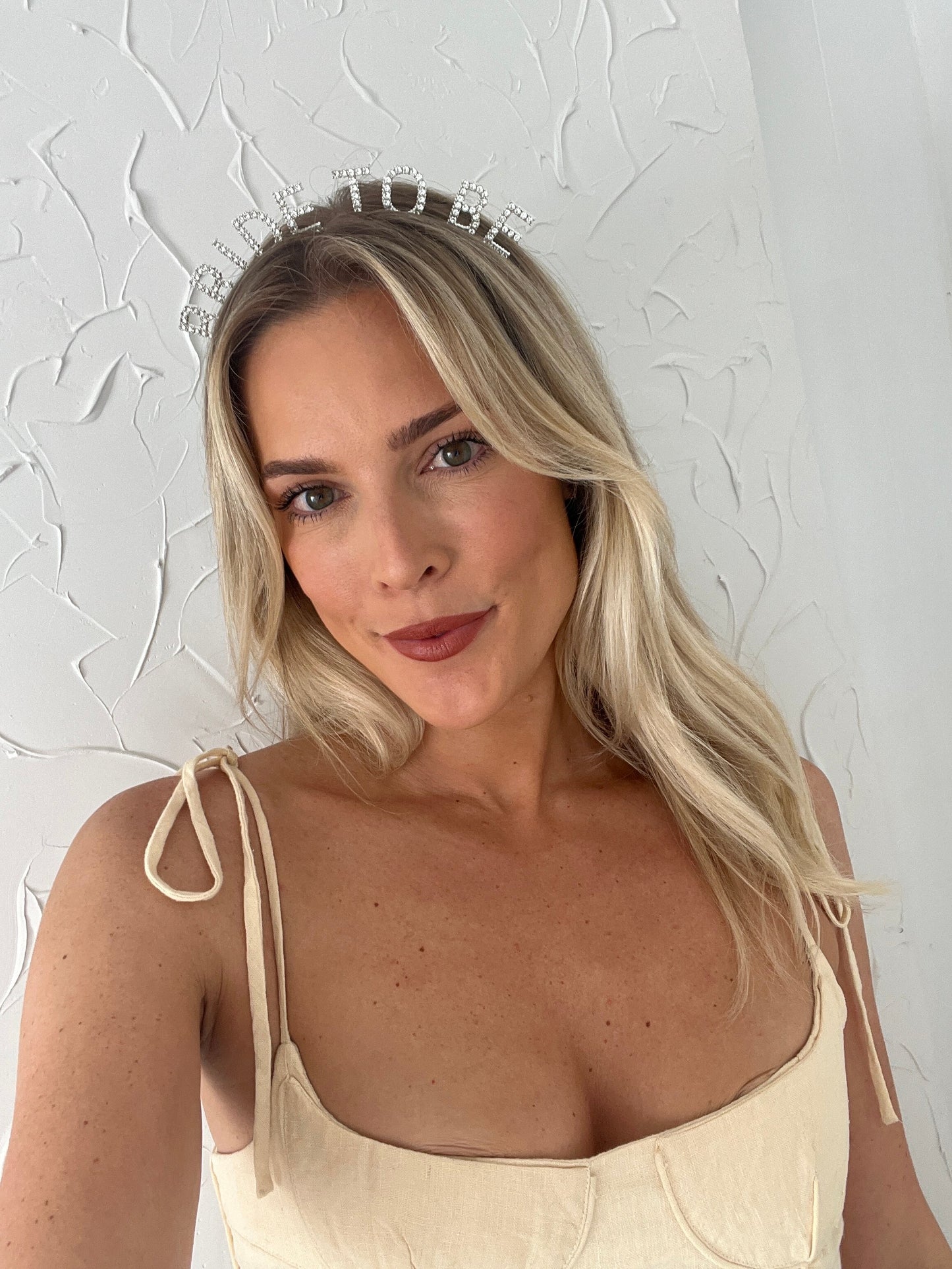 A woman wearing our 'Bride-to-be' Diamante Headband against a textured neutral wall. The silver headband lettering is detailed with diamantes, Adding elegance and sparkle to any bride for pre wedding celebrations or hen party.