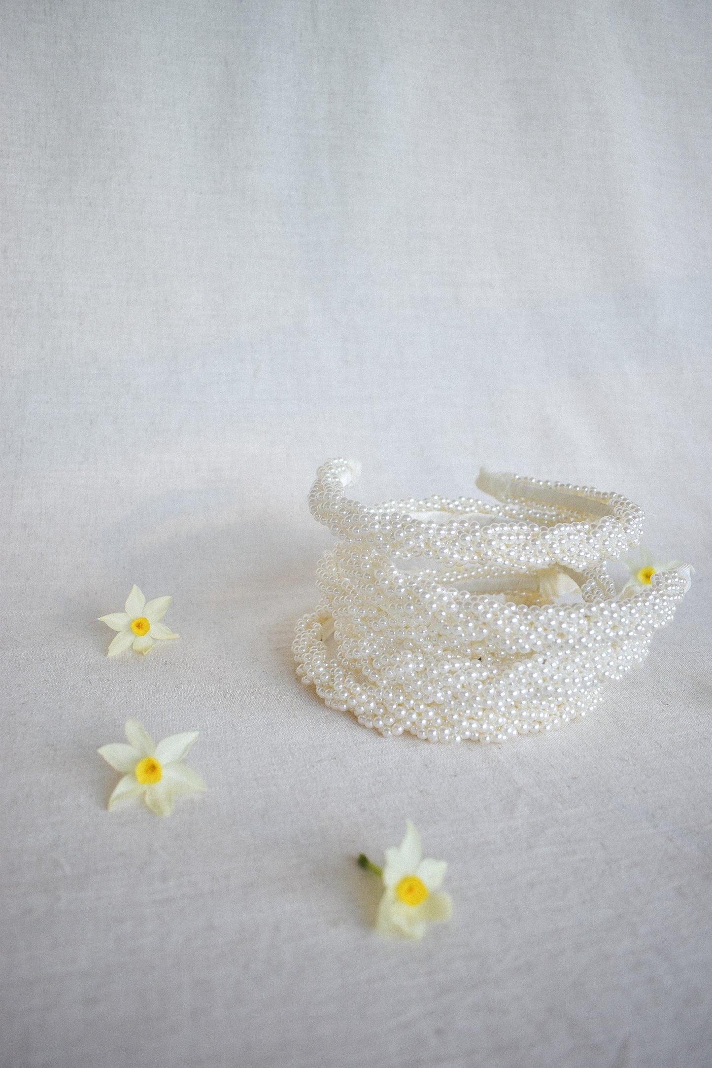 A stack of our Buttercup Headband on a textured fabric surface, complimented by a few small white and yellow flowers, laying on their side. Showing the detail of the small pearl braided design end to end of the band. Perfect for enhancing a day or evening outfit at your next event or spring season.