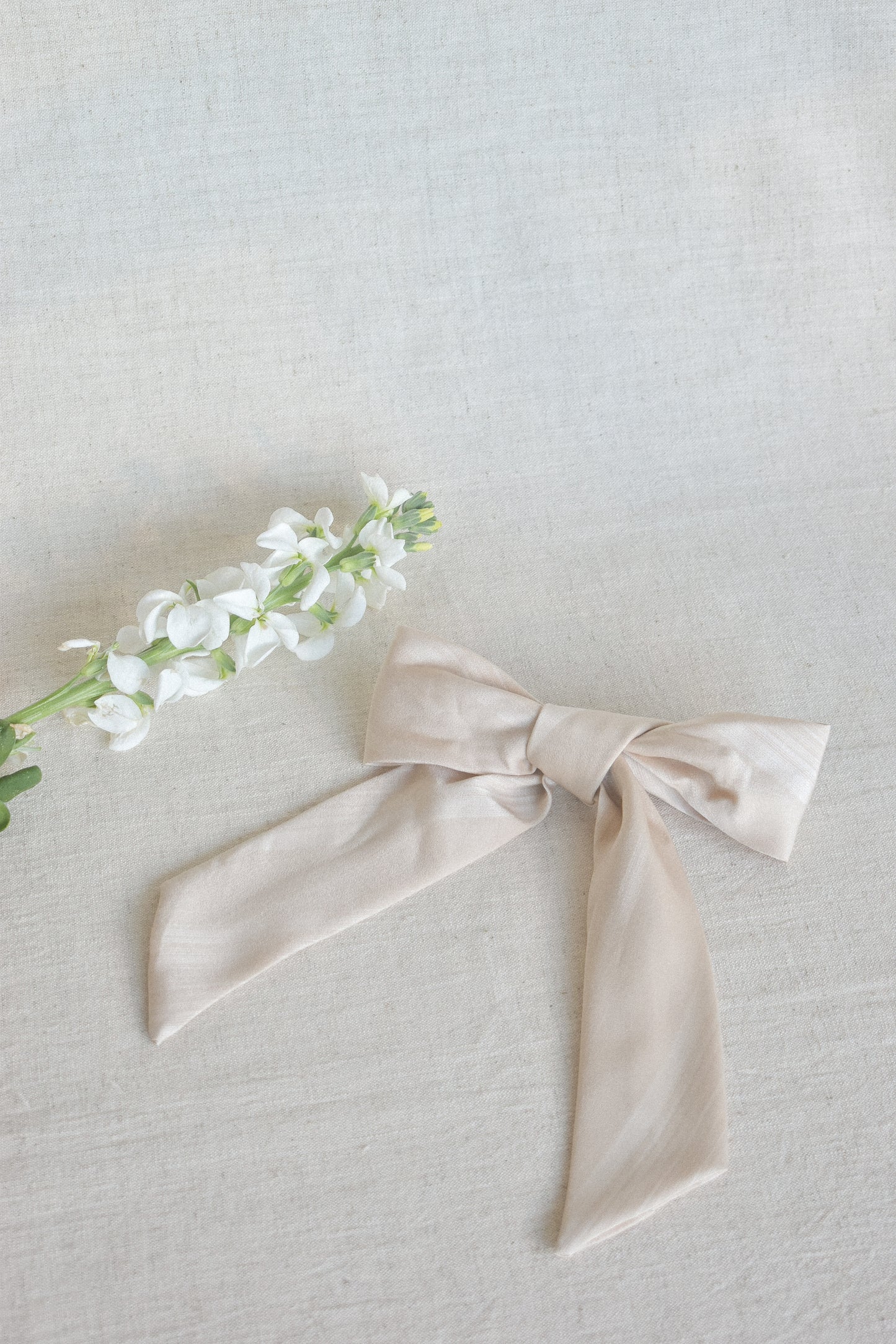 Our Blossom Bow Hair Clip, laying flat next to a white flower complimenting the creamy latte colour with a satin/silk material. Perfect for brides or bridesmaids