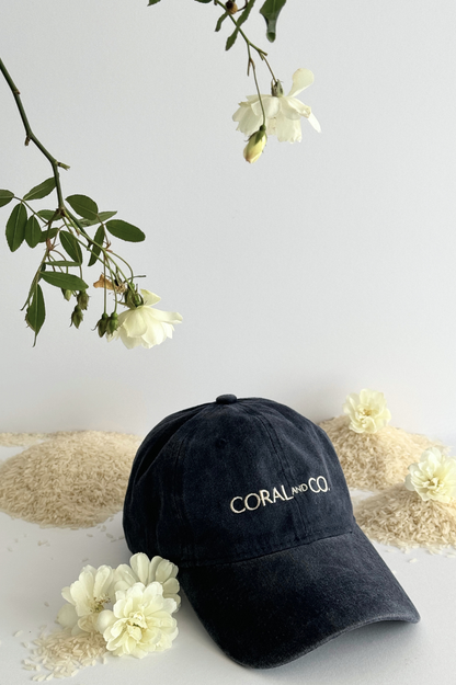 Our Blueberry Pie Cap placed on a white surface with cream flowers compliments the faded blueberry-colour denim cap with cream embroidery reading, CORAL AND CO, at the front. It's the perfect accessory for day to day and adding a pop of colour to any outfit