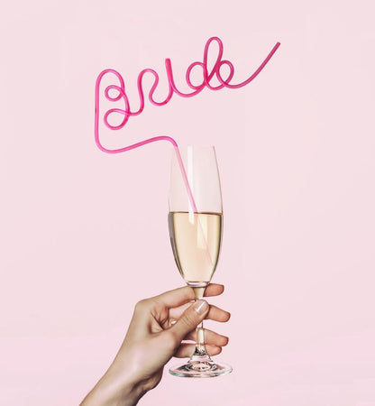 Our pink BRIDE straw being used in a glass of Champagne, with a light pink background. Highlighting the height and colour of the straw and the large BRIDE lettering. Functional with all beverages on any hen/bachelorette party. Fun gift for the bride to be