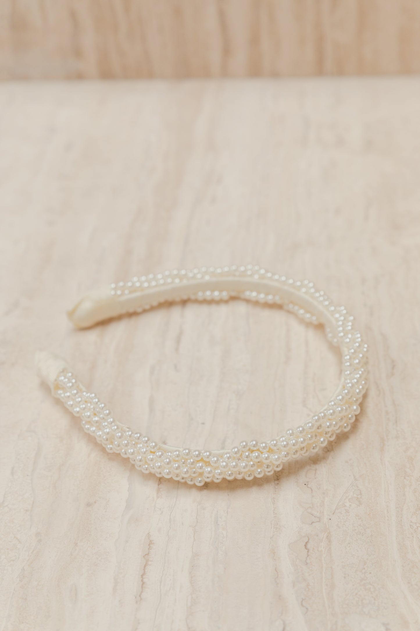 Our Buttercup Headband on a neutral surface, laying on it's side. Showing the small pearl braided design end to end of the band. Perfect for enhancing a day or evening outfit at your next event.