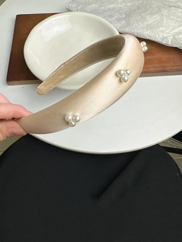 Our Champagne coloured satin padded headband with small clustered pearl detail across the band. Band is held on an angle against a table set up, highlighting the satin sheen and colour of the padded band. Add a touch of glamour to any outfit day or night. Perfect for bridal events or date night