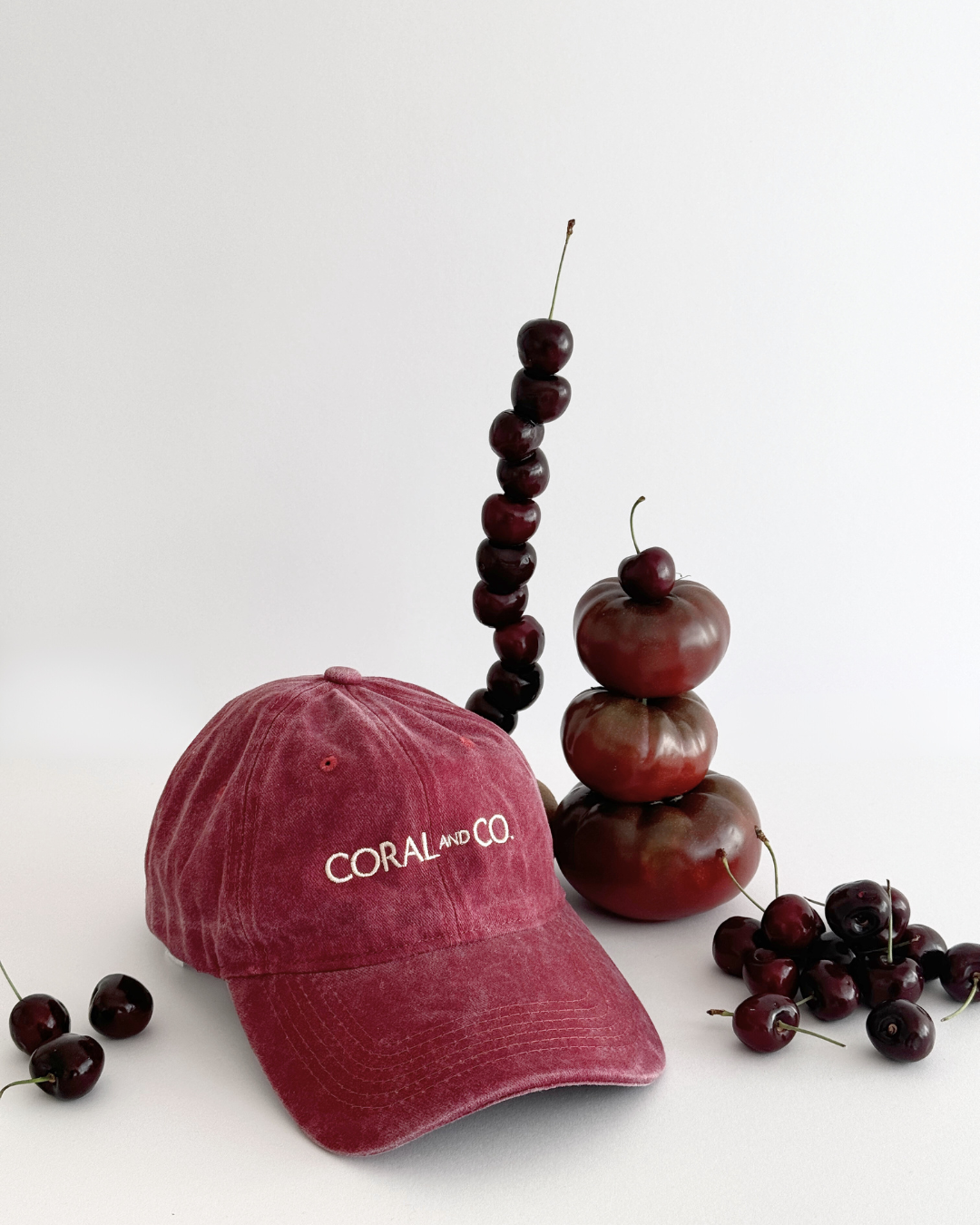 Our Cherry glaze, burgundy/maroon cap, faded denim material with a cream embroidery reading, CORAL AND CO. Cap is sitting against a white background with cherries scattered, showcasing the gorgeous colour of this cap. Perfect for day to day, adding a cool casual pop of colour to any outfit