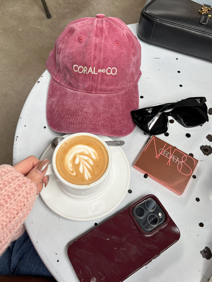 Our Cherry glaze, burgundy/maroon cap, faded denim material with a cream embroidery reading, CORAL AND CO. A birds eye view of a table set up next to a coffee, make up palate, smart phone and sunglasses, offering a size comparison. Perfect for day to day coffee catch ups, adding a cool casual pop of colour to any outfit