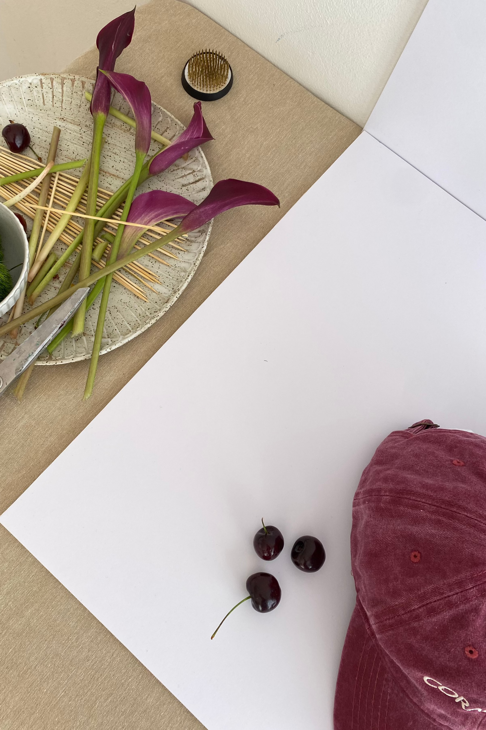 Our Cherry glaze, burgundy/maroon cap, faded denim material with a cream embroidery reading, CORAL AND CO. Birds eye view of our cap sitting against a white background with cherries scattered and maroon flowers, highlighting the gorgeous colour of this cap. Perfect for and casual or sport occasion on a sunny day