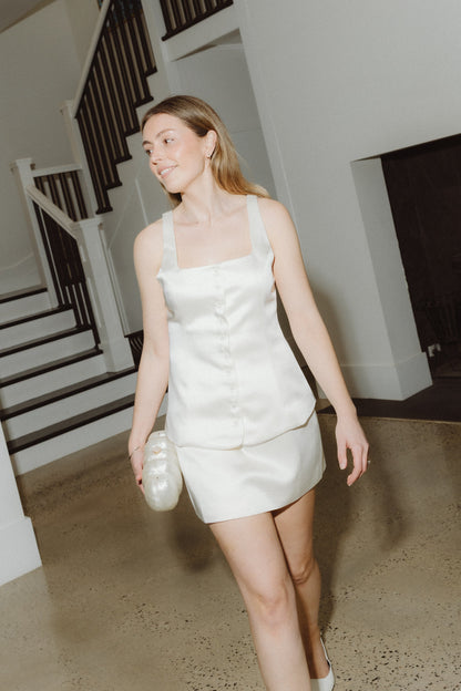 Woman in a white dress standing in a room with a staircase, holding the pearl cloud clutch bag.