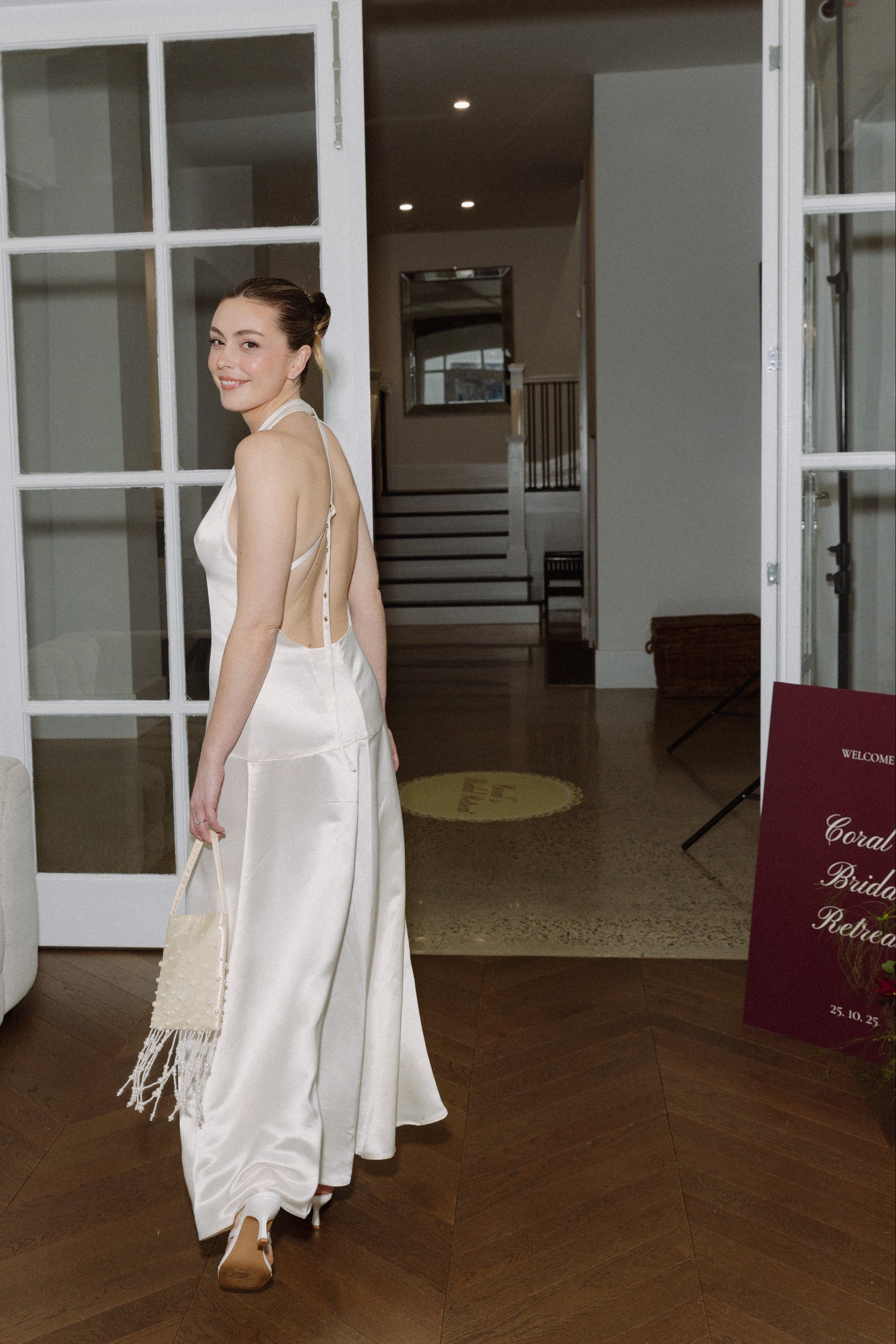 Woman in a white dress with a backless design standing in a room with wooden flooring and large glass doors, holding an elegant satin evening bag with pearls and fringe details