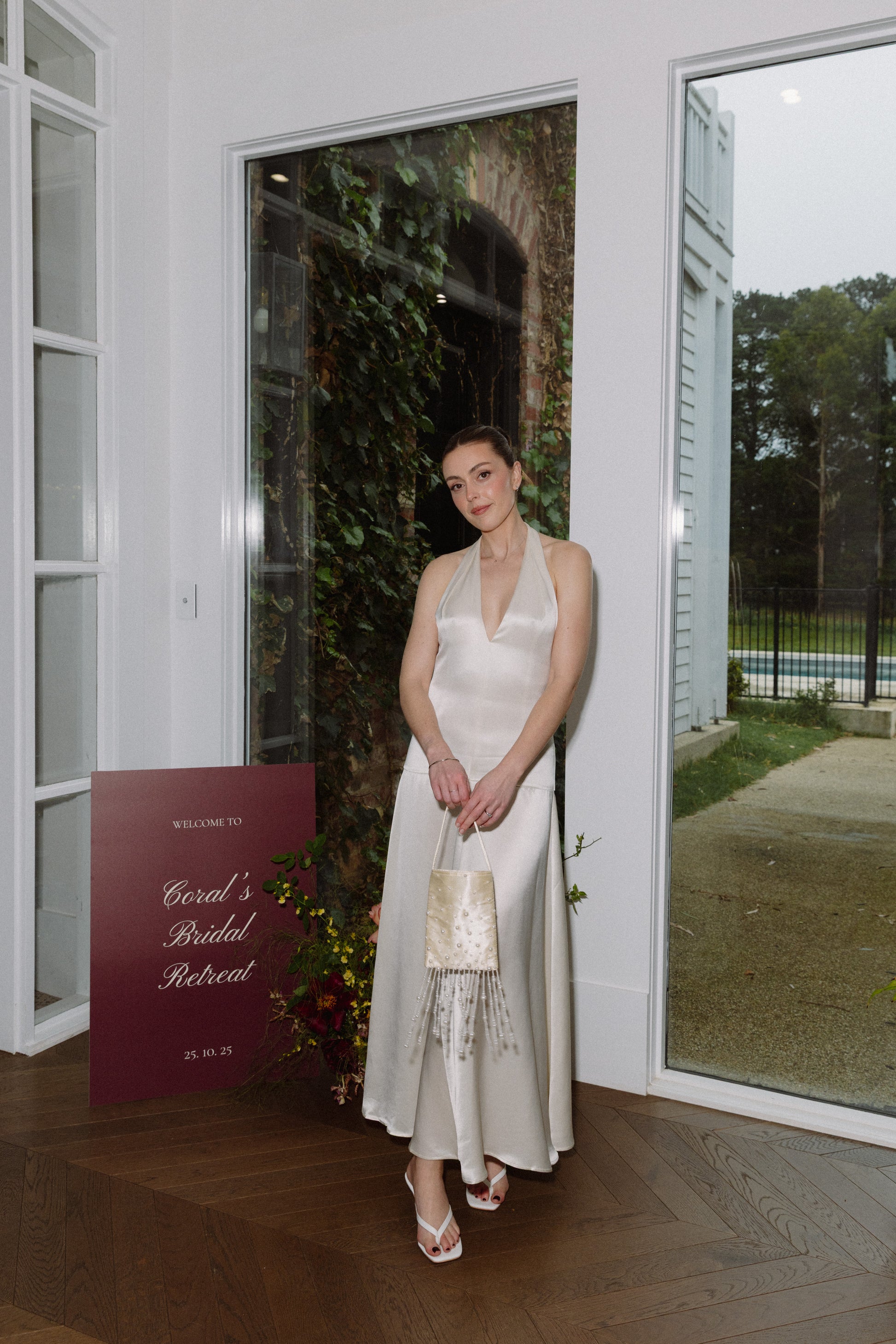Woman in a white dress standing in a room with large windows, holding a small bag, the Goldie Bag featuring delicate pearls and tassel beads, perfect for weddings and events.
