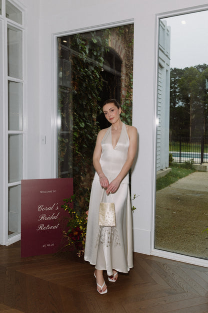 Woman in a white dress standing in a room with large windows, holding a small bag, the Goldie Bag featuring delicate pearls and tassel beads, perfect for weddings and events.