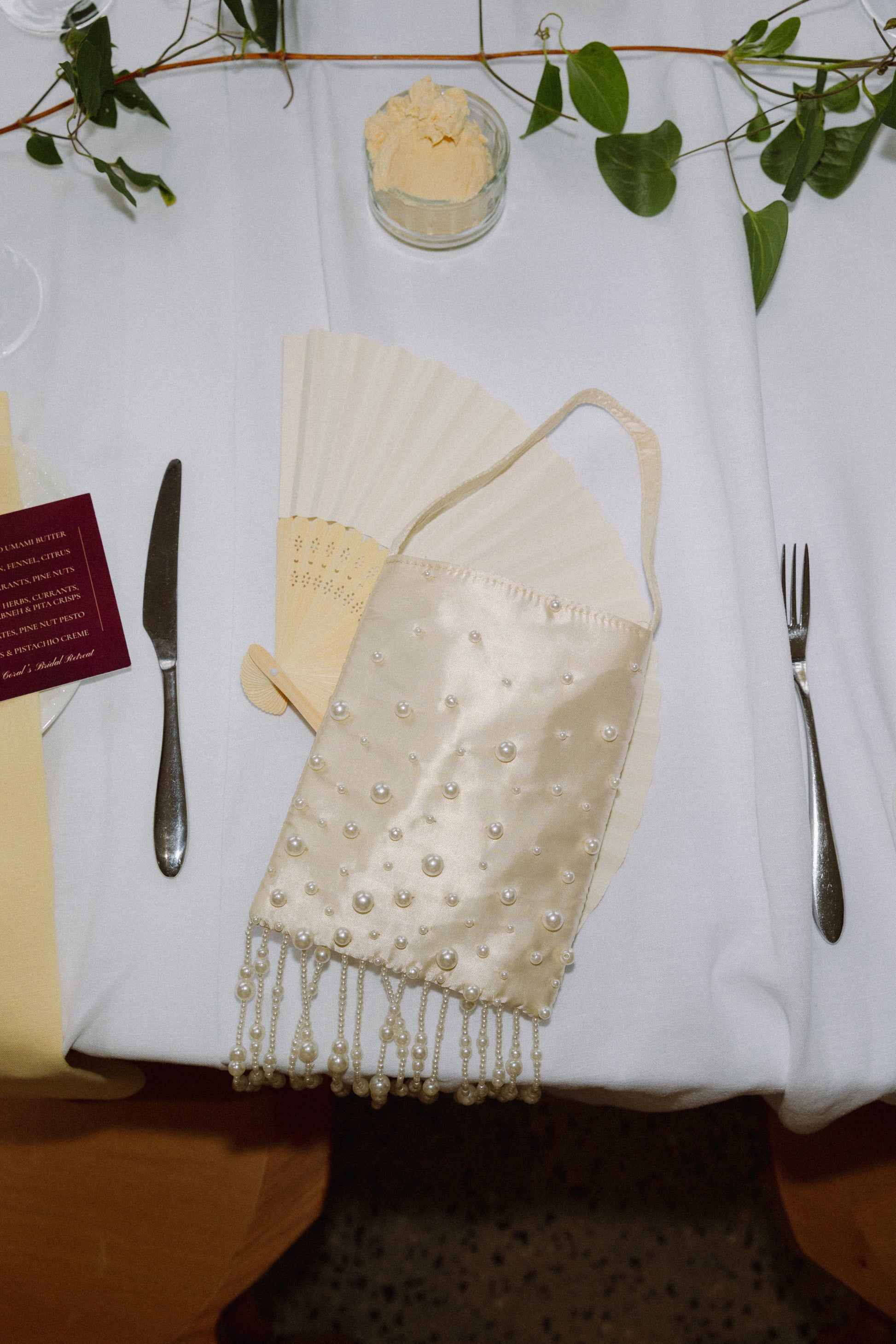 Decorative table setting with a embroidered napkin, knife, and fork on a white tablecloth. Laying on top is a Gold satin pearl-embellished mini bag with beaded tassel detail by CORAL & CO.