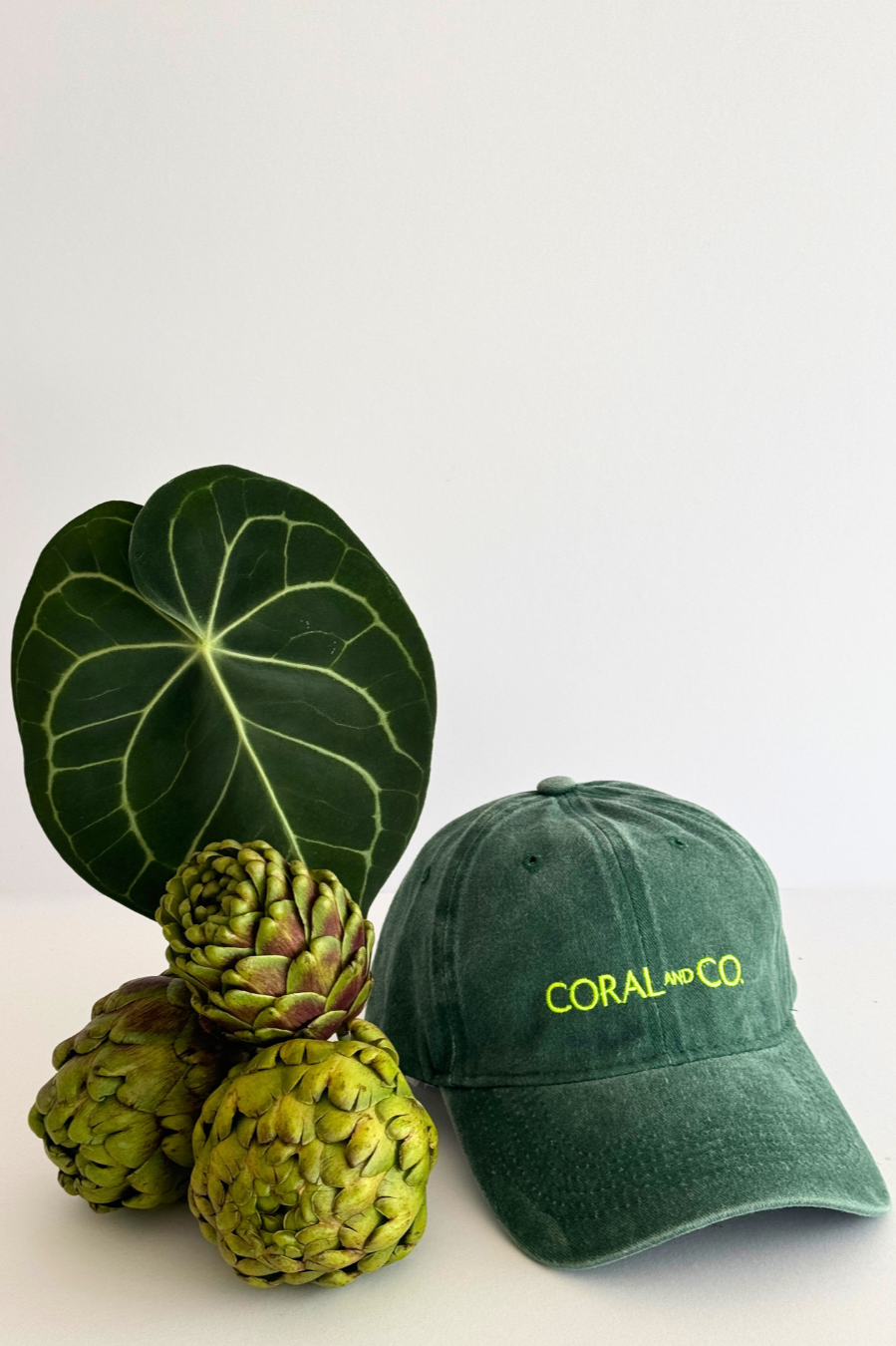 Our Green Goddess Cap, on a white surface next to a green flower arrangement. Dark green faded denim and featuring a bold fluro green embroidery. Perfect for casual daily wear