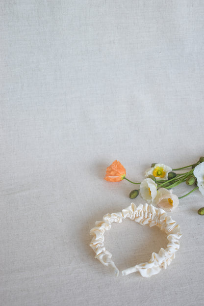 Out Lily soft satin headband show here laying flat with spring flowers around. Highlighting the ruffled design and sheen from the cream satin/silk material. Neutral colour that will compliment any outfit, especially bridal