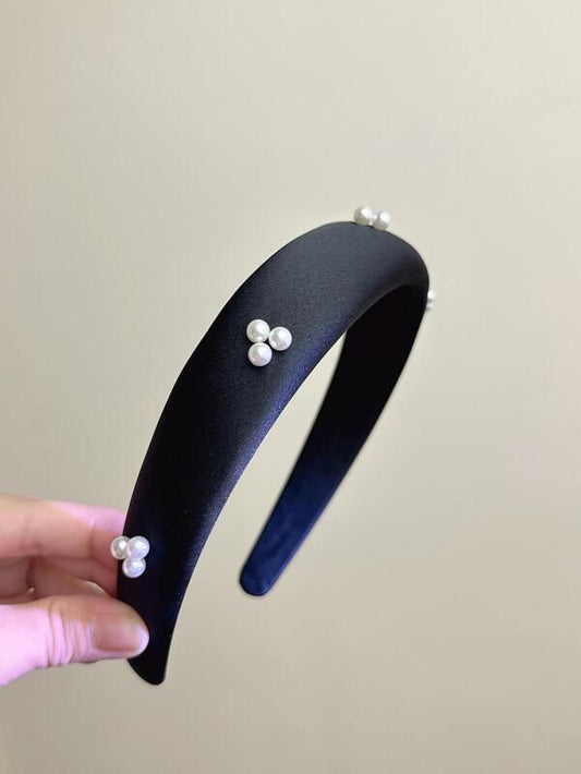 Our Noir headband being held at an angle, showing the sheen of the black satin fabric and the small pearls clustered and spaced out along the band. Highlighting the width of the band design. Perfect for formal events