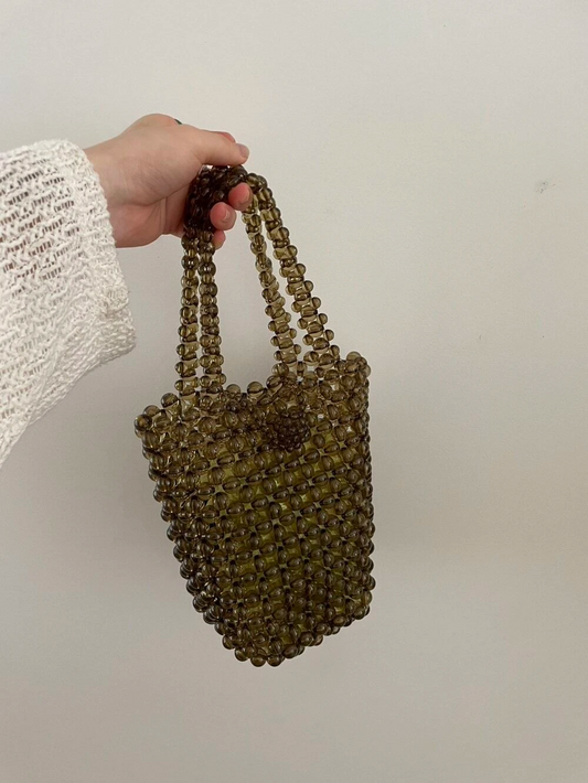 Our Olive Beaded Bucket Bag being held against a white background. Olive khaki clear beaded bag with a loop over to close clasp and a short over the forearm beaded handle. Effortlessly elevates your party outfit