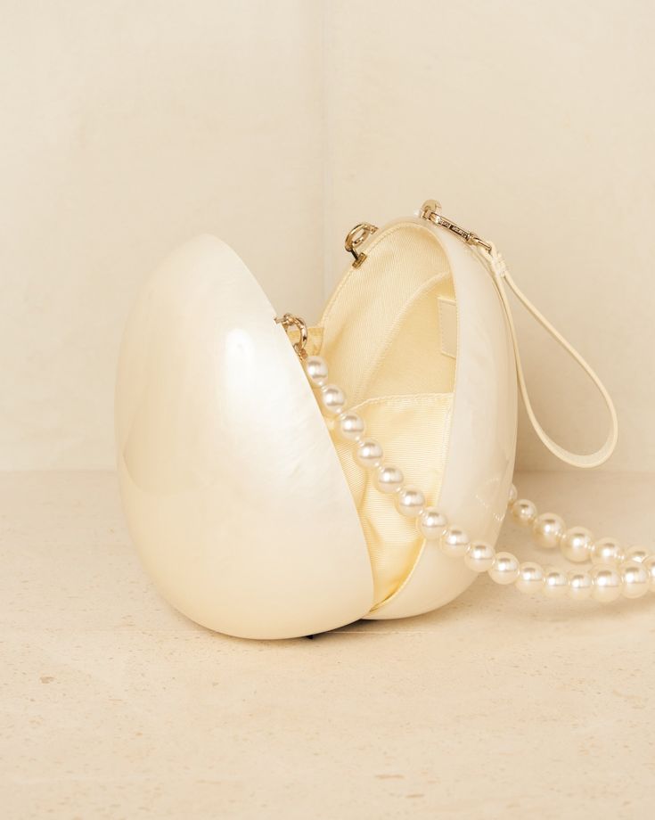 A close-up of our Peggy Bag, a white acrylic egg-shape, shown against a neutral background, open and from the side. Highlighting the space within the bag and inner silk sleeves that stop contents falling out as well as the sheen from the acrylic and the pearls. Shown with both handle attachments. The ultimate bridal accessory or statement event bag whilst holding you make up essentials