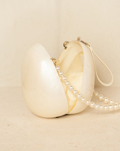 A close-up of our Peggy Bag, a white acrylic egg-shape, shown against a neutral background, open and from the side. Highlighting the space within the bag and inner silk sleeves that stop contents falling out as well as the sheen from the acrylic and the pearls. Shown with both handle attachments. The ultimate bridal accessory or statement event bag whilst holding you make up essentials