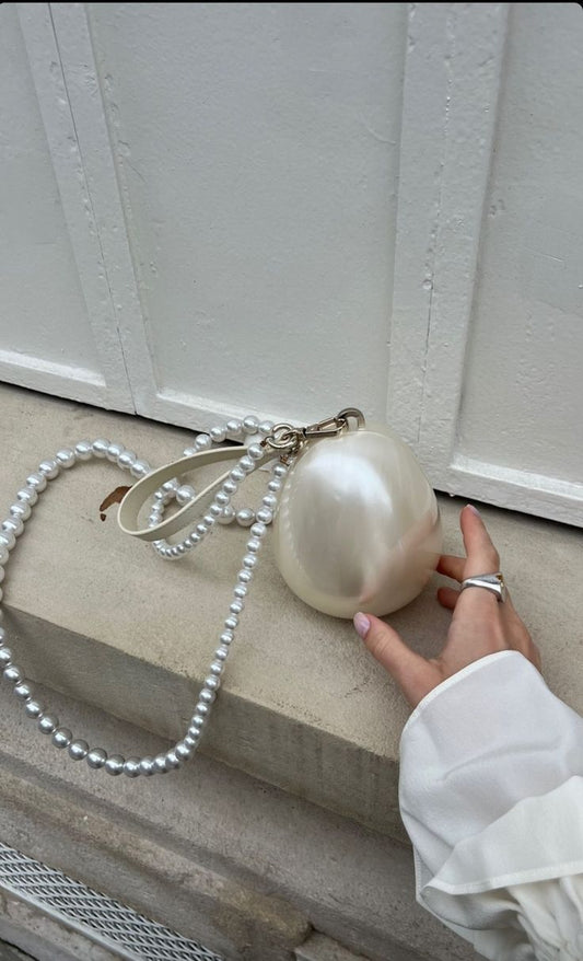 Our Peggy Bag, a white acrylic egg-shaped mini bag, shown against a hand for size comparison. Versatile interchangeable handles, as a clutch with a small leather wrist strap or a cross body pearl beaded shoulder bag. The ultimate bridal accessory or statement event bag. 