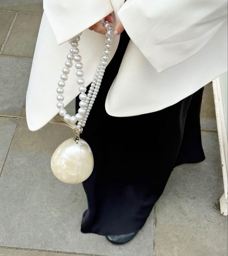 Our Peggy Bag being worn by a woman wearing black and white, highlighting the cream sheen of the egg-shaped acrylic bag. The woman is holding the bag using the longer pearl handle. Adding a fun and classy moment to any formal evening event