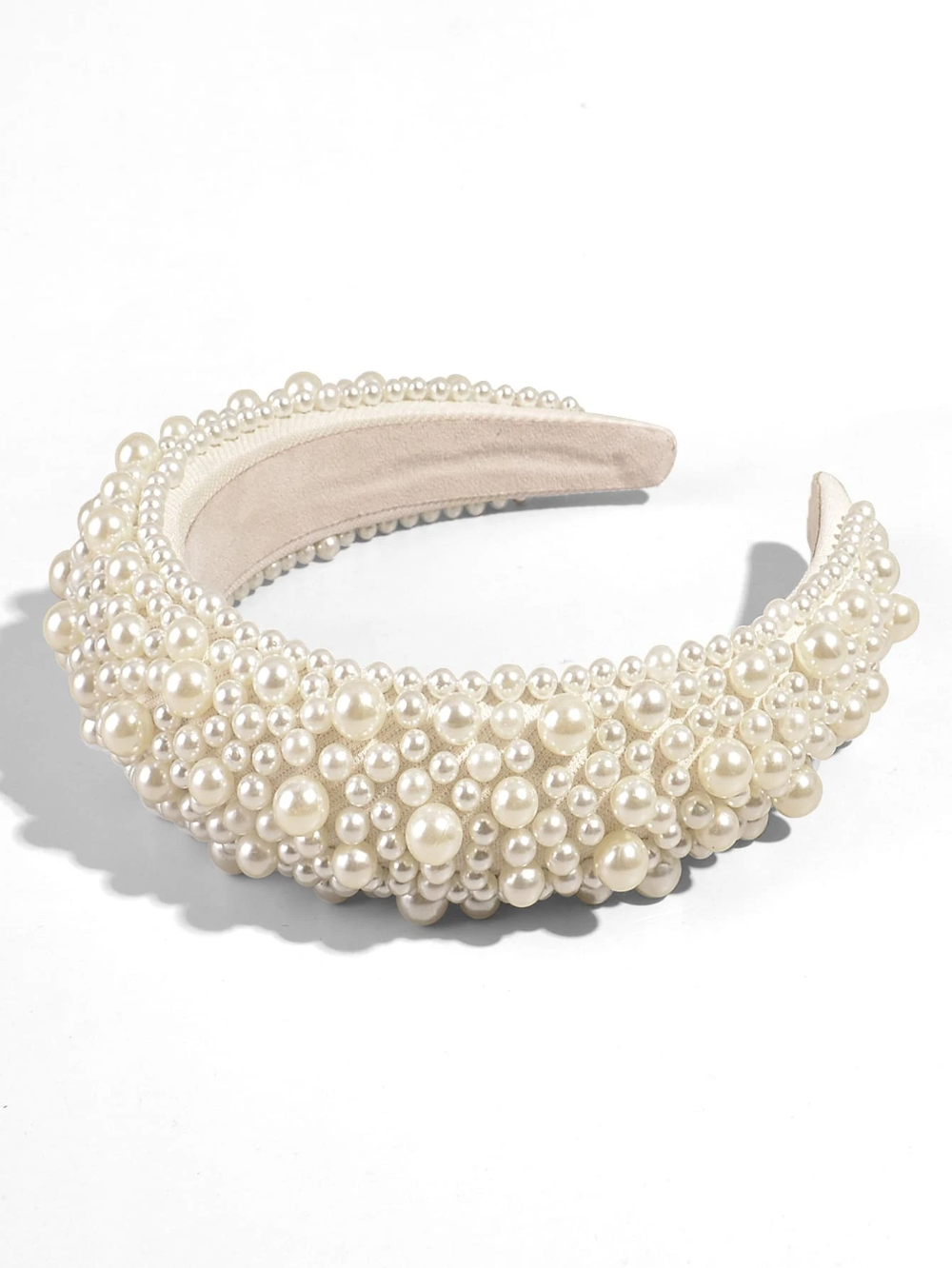 Our Peony Headband laying flat on a white surface, showing the multiple sizes of faux pearls across the padded cream band. Highlighting the sheen of the pearls and angle shows the height of the padded band. Perfect bride or bridesmaid accessory or daytime event