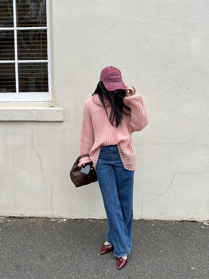 A woman wearing our Cherry glaze, burgundy/maroon cap, faded denim material with a cream embroidery reading, CORAL AND CO. She is stood against a white wall with casual jeans and pink knit jumper, inspiring how you could style this cap. Perfect for day to day, adding a cool casual pop of colour to any outfit