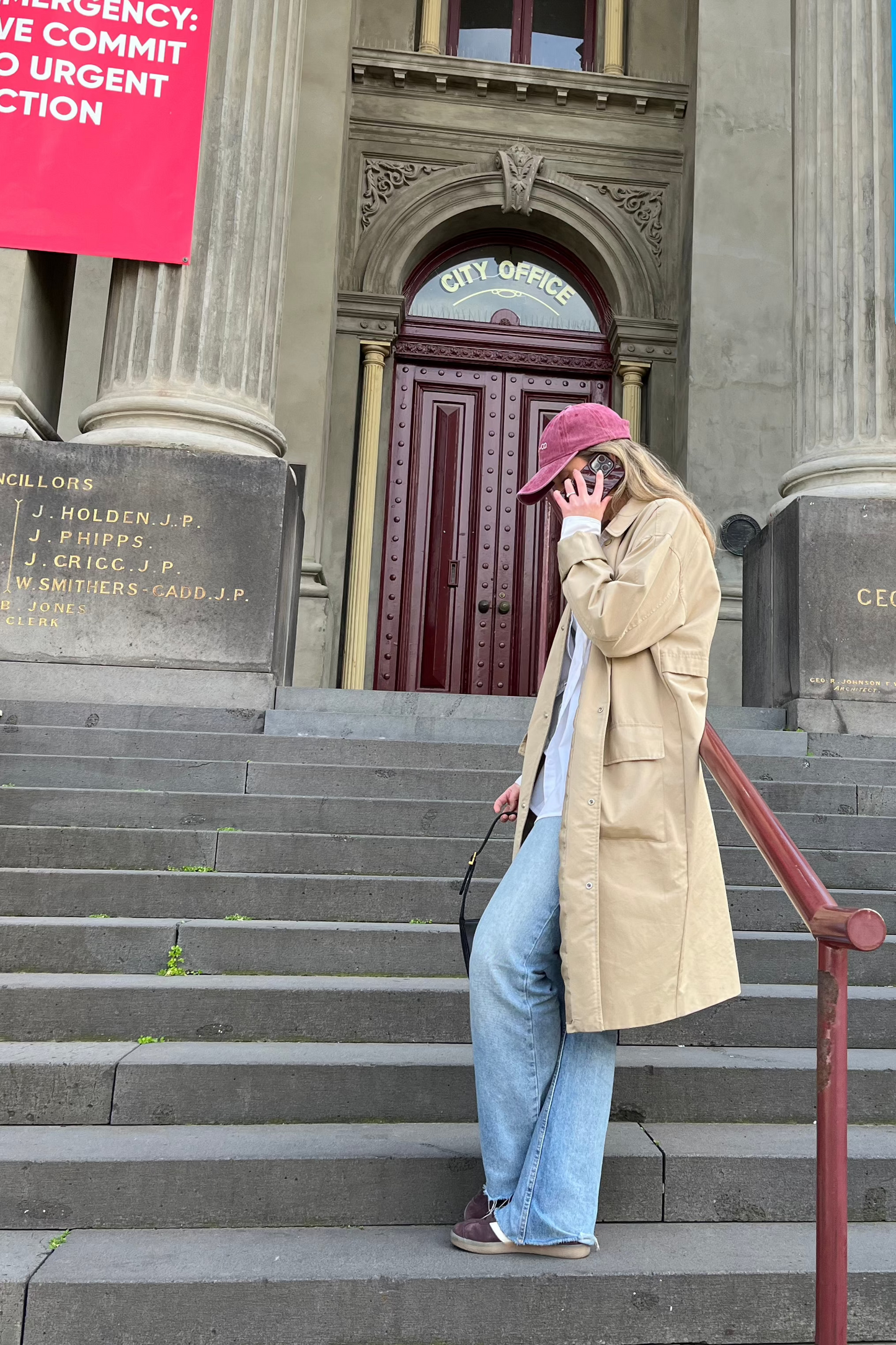 A woman wearing our Cherry glaze, burgundy/maroon cap, faded denim material with a cream embroidery reading, CORAL AND CO. Stood to the side, highlighting the profile angle of our cap. She is stood on a concrete staircase with a parker coat on, showcasing the gorgeous cap worn whilst running errands or shopping