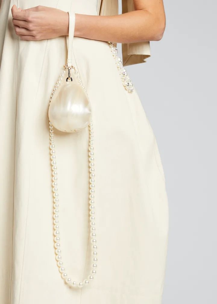 Our Peggy Bag being worn by a woman wearing a cream dress, highlighting the cream sheen of the egg-shaped acrylic bag. The woman is holding the bag using the short cream leather wrist strap and the longer pearl handle is dangling down, adding more class and glamour to any bridal party