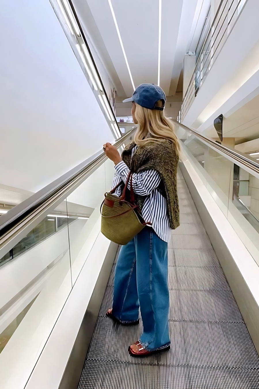 A blonde woman, stood on an escalator wearing our faded denim Blueberry Pie Cap. Showing more of the back of the cap which is adjustable. She's wearing blue denim jeans and an oversized shirt. The cap adds a cool fun element to her day outfit