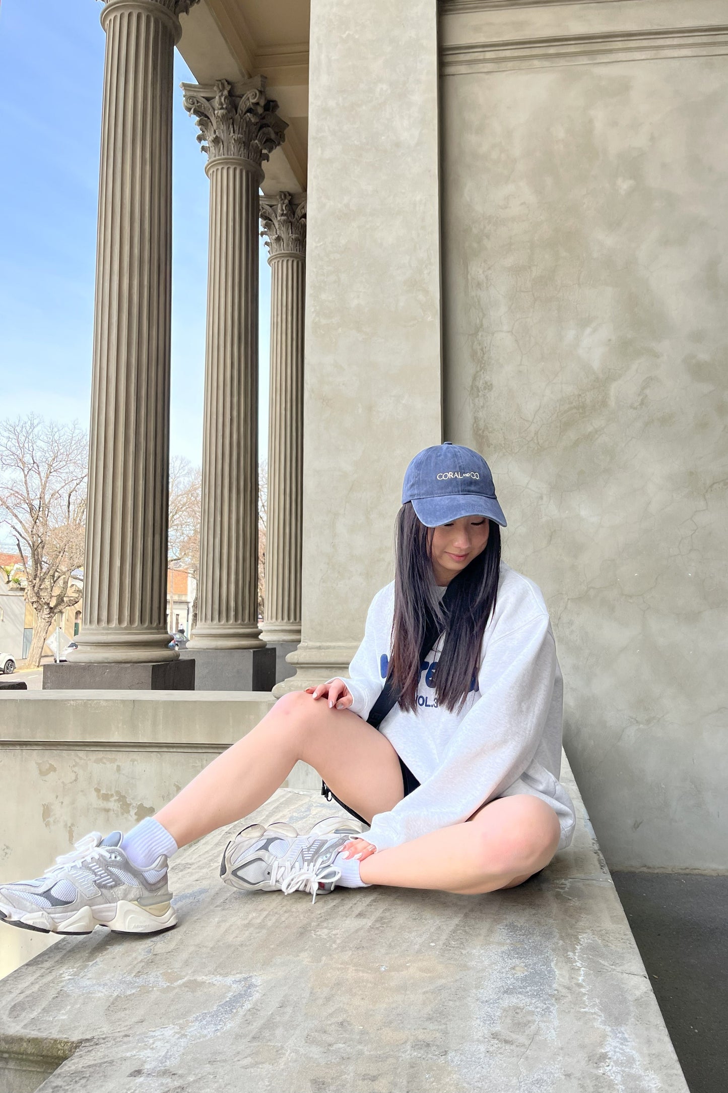 A girl sat on a marble surface on holiday, wearing our faded blueberry-coloured denim cap with cream embroidery reading, CORAL AND CO, at the front. Perfect holiday accessory or sporty casual outfit.