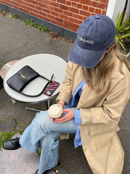 A woman sat on a chair, next to a table wearing our faded blueberry-coloured denim cap with cream embroidery reading, CORAL AND CO, at the front. It's the perfect accessory for day to day activities, your next holiday or sporting events